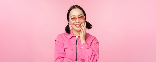 Close up ritratto di elegante donna asiatica in occhiali da sole sorridente guardando lusingato ridendo civettuola in piedi su sfondo rosa