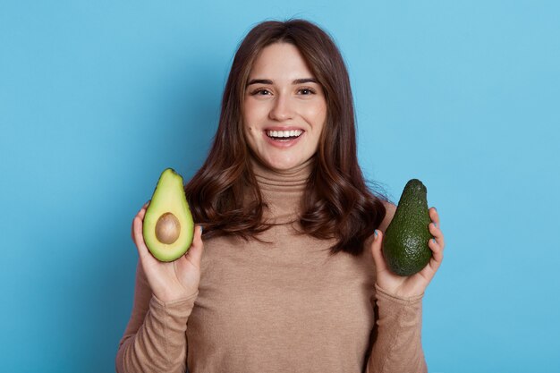 Close up ritratto di giovane donna dai capelli scuri con due metà di avocado fresco verde in posa isolato sopra il muro blu, donna sorridente con i capelli scuri preferisce il cibo biologico.
