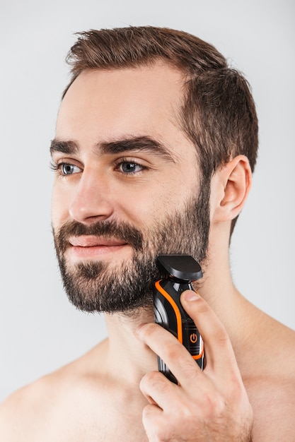 Close up ritratto di un bell'uomo barbuto che si rade con un rasoio elettrico isolato su bianco