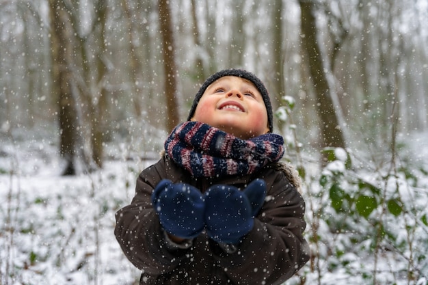 Close up ritratto di un ragazzino che gioca con i fiocchi di neve in un parco in inverno. Il bambino felice gode della prima neve in una foresta.