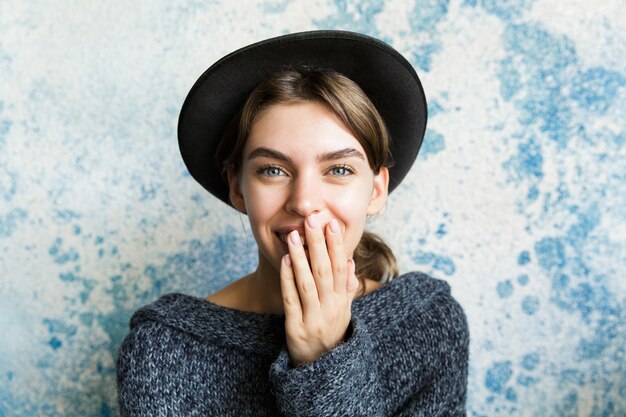 Close up ritratto di una giovane donna vestita in maglione e cappello oltre il muro blu, ridendo, coprire la bocca