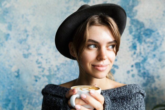 Close up ritratto di una giovane donna vestita in maglione e cappello oltre il muro blu, tenendo la tazza di caffè caldo, guardando lontano