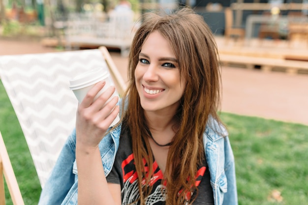 Close up ritratto di una ragazza sorridente con lunghi capelli scuri e un sorriso affascinante vestito con una giacca di jeans. Sta guardando la fotocamera con un grande sorriso aperto e tiene una tazza di caffè. Bella ragazza nel parco