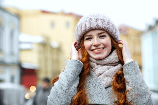 Closeup ritratto di allegra donna dai capelli rossi che indossa berretto caldo lavorato a maglia e sciarpa in posa per la strada. Spazio per il testo