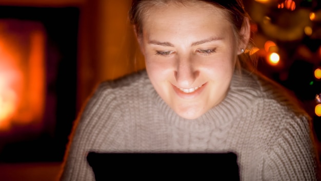 Closeup ritratto di donna sorridente felice con tavoletta digitale seduta accanto al caminetto di notte