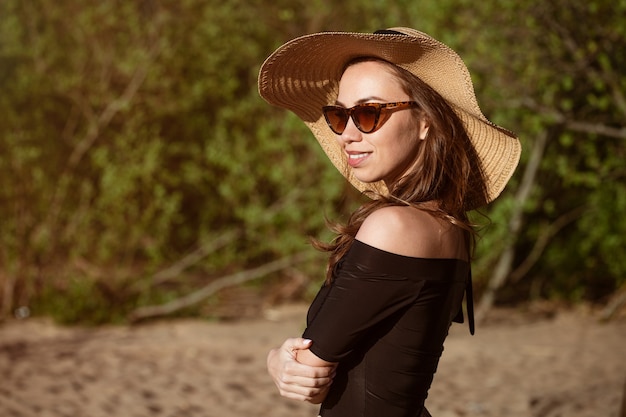 Closeup ritratto di una donna in un cappello dal sole in una giornata di sole estivo bella giovane donna caucasica in posa in occhiali da sole all'aperto nel pomeriggio carino sorridente