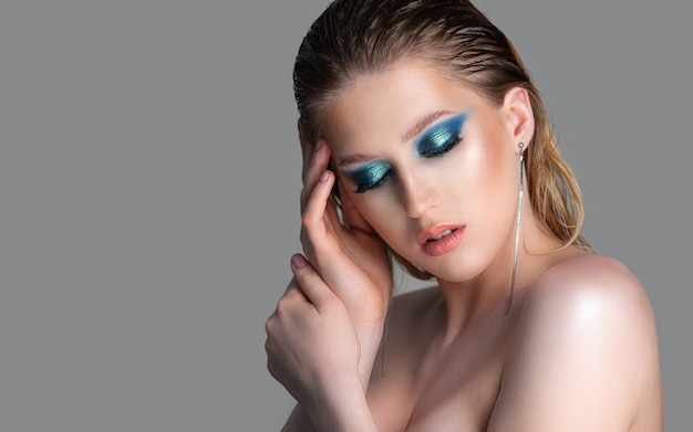 Closeup ritratto in studio di un'elegante donna bionda con i capelli bagnati e trucco occhi smokey blu profondo. Modello in posa con le spalle nude su uno sfondo grigio. Spazio vuoto