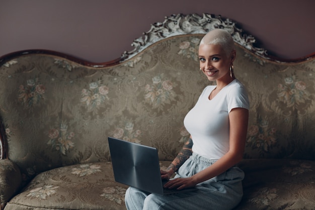 Coaching giovane donna con corti capelli biondi ritratto seduto e sorridente con il computer portatile sul divano vintage