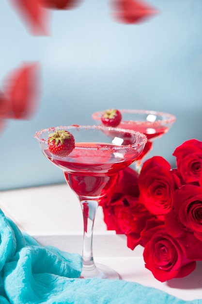 Cocktail alcolico esotico rosso in bicchieri trasparenti e petali di rose rosse sul tavolo bianco in legno per una cena romantica.