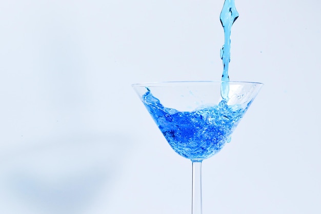 Cocktail con liquido blu in vetro. Vetro con acqua blu versando con liquido con spruzzi e gocce. Bicchiere da Martini riempimento con alcool con schizzi su sfondo bianco. Bevanda rinfrescante concetto.