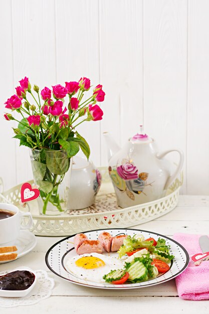 Colazione a San Valentino - uovo fritto a forma di cuore, toast, salsiccia e verdure fresche. Tazza di caffè. colazione inglese