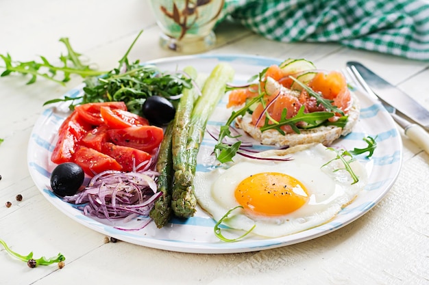 Colazione chetogenica Colazione Pagnotte croccanti all'uovo fritto con asparagi verdi di salmone pomodori freschi e cipolle rosse su piatto bianco