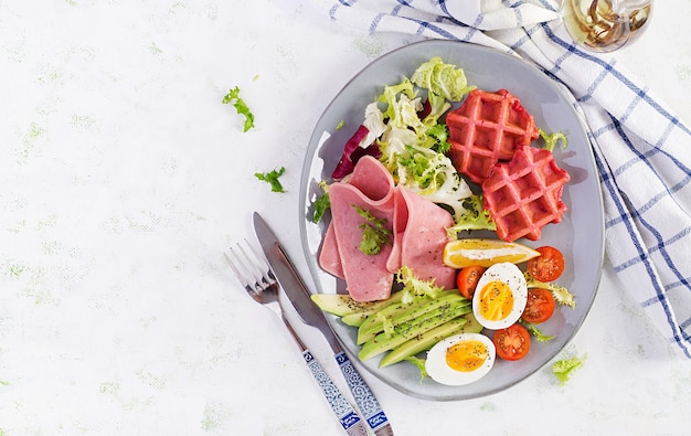 Colazione con waffle di barbabietola, uovo sodo, prosciutto, pomodoro e fetta di avocado su superficie bianca