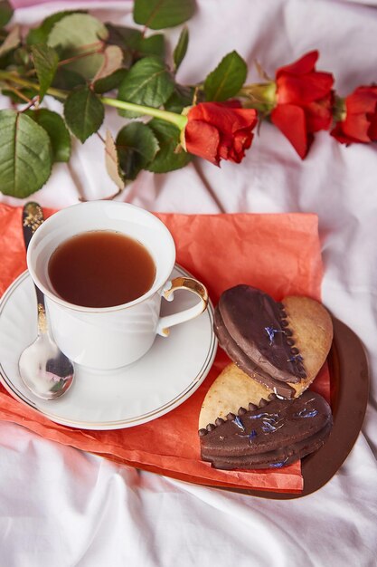 Colazione di San Valentino a letto tra rose rosse tradizionali biscotti vegani a forma di cuore e tazza di caffè sulle lenzuola Estetica romantico sfondo vacanza lagom