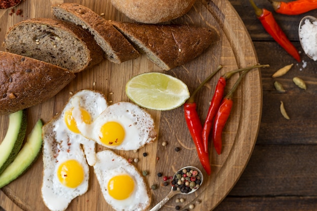 Colazione di uova fritte, pane, avocado e spezie varie su una tavola di legno.