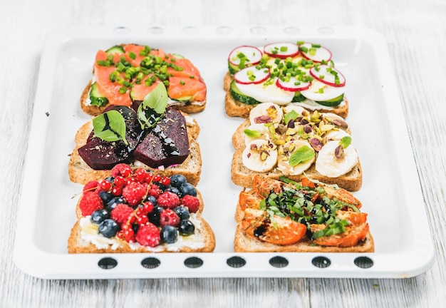 Colazione dolce e salata toast varietà panini con frutta verdura uova salmone affumicato su teglia bianca su sfondo di legno chiaro