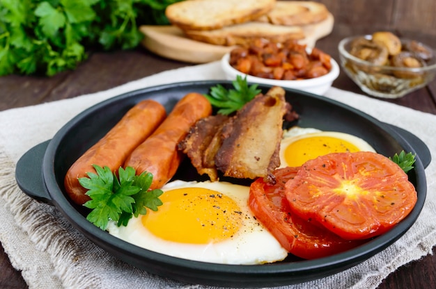 Colazione inglese: salsicce, pancetta, pomodori, uova, fagioli in salsa, funghi fritti, toast su un tavolo di legno scuro. Vista dall'alto. Cibo classico tradizionale dell'Inghilterra.