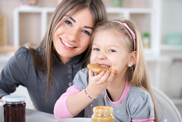 Colazione madre e figlia in cucina. La bambina sveglia mangia il pane con il burro di arachidi. Guardando la fotocamera.