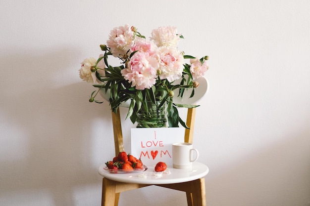 Colazione per la festa della mamma Piatto bianco a forma di cuore con fragole fresche regalo tazza di caffè e bouquet di peonie con regalo a letto Composizione di natura morta Buona festa della mamma