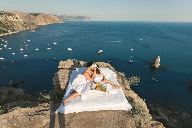 Colazione romantica di un uomo e una donna in un letto bianco su una scogliera a picco sul mare e sugli yacht