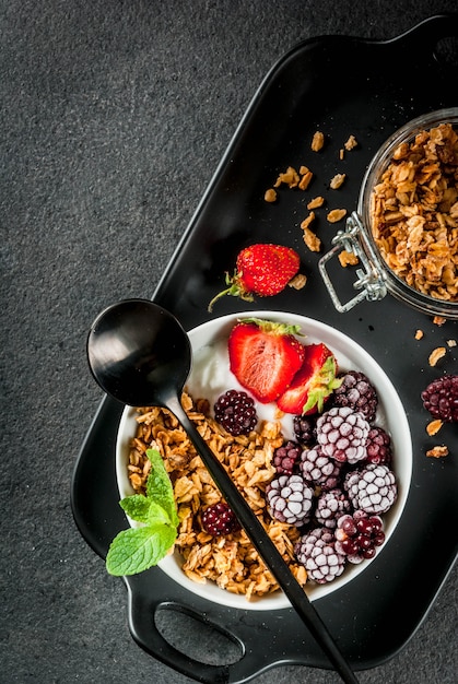 Colazione salutare. Frutti e bacche estive. Yogurt greco fatto in casa con muesli, more, fragole e menta. tavolo in pietra nera, con gli ingredienti. Vista dall'alto