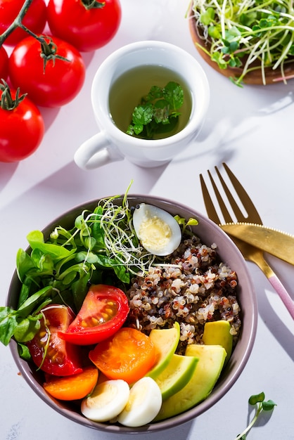 Colazione vegetariana con quinoa, uova, microgreen e verdure, tè verde alla menta su un tavolo bianco, piatto lay