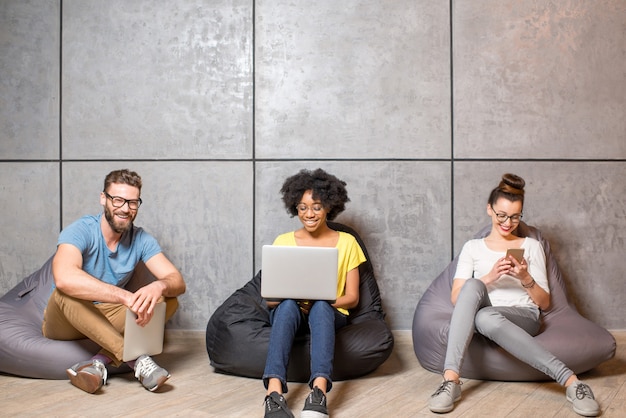 Collaboratori multietnici vestiti casualmente con abiti colorati che lavorano con diversi gadget seduti sulle sedie della borsa sullo sfondo del muro grigio