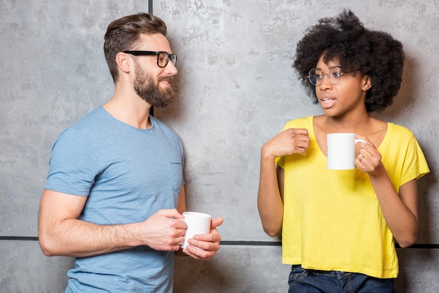 Colleghe multietniche vestite casualmente per una pausa caffè vicino al muro grigio al chiuso