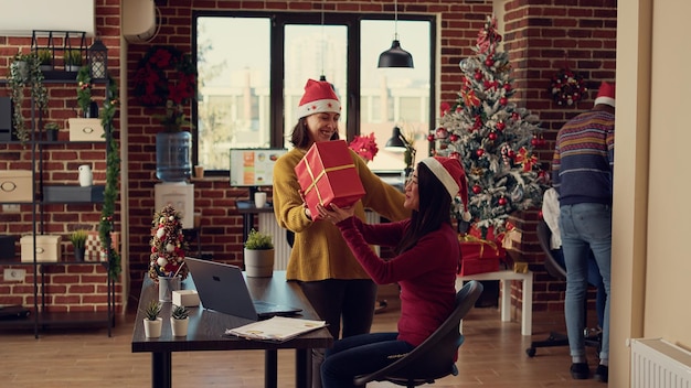 Colleghi che fanno regali per celebrare la vigilia di natale al lavoro, festeggiano le vacanze invernali in ufficio con albero di natale e decorazioni. Diverse donne che si scambiano doni durante le feste.