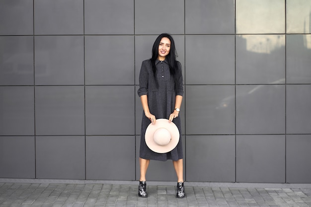 Collezione autunno alla moda. Bella ragazza felice del brunette che porta vestito grigio alla moda che tiene cappello alla moda nelle mani. Modello in abito alla moda in posa sul muro grigio. Ritratto all'aperto.