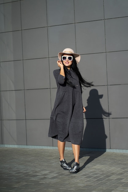 Collezione primavera alla moda. Bella ragazza felice del brunette che porta vestito grigio alla moda e cappello alla moda. Modello in abito alla moda in posa sul muro grigio