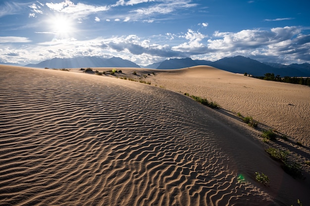 Colline sabbiose illuminate dal sole ondeggiano dal vento con vegetazione desertica sullo sfondo della catena montuosa di Kadar e un cielo leggermente nuvoloso