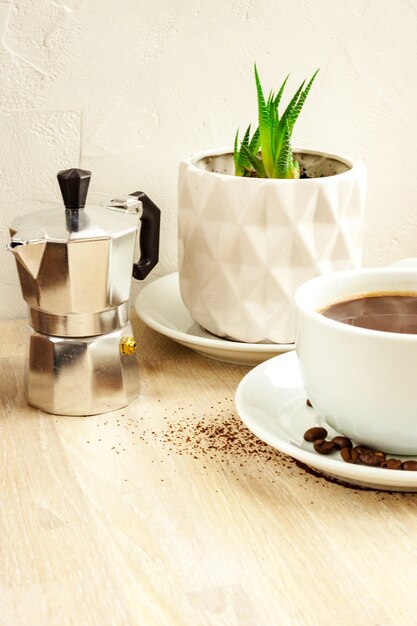 Colore bianco Tazza di caffè nero con piattino e fagioli, caffettiera in alluminio, pianta grassa verde in vaso bianco sulla tavola di legno beige. Messa a fuoco selettiva. Copia spazio