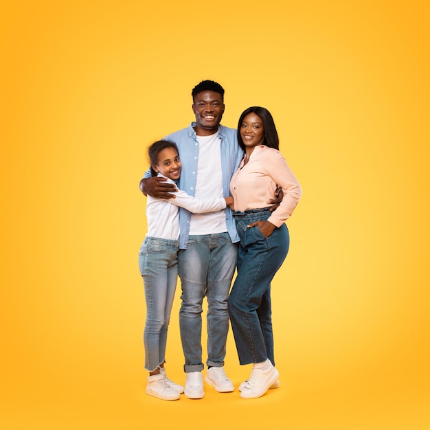 Colpo a figura intera di felice famiglia afroamericana che abbraccia e sorride alla telecamera su sfondo giallo