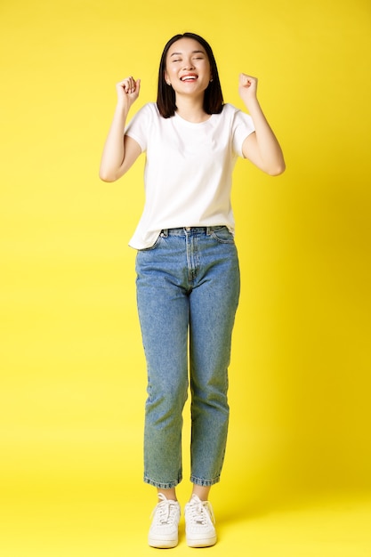 Colpo a grandezza naturale di donna asiatica allegra in jeans e maglietta bianca che salta dalla felicità, vincendo il premio e gioendo, celebrando e gridando sì con gioia, sfondo giallo.