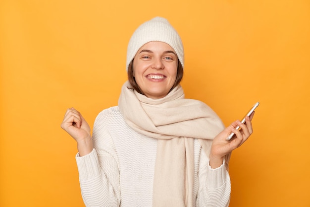 Colpo al coperto di attraente sorridente felice donna caucasica che indossa un berretto caldo e una sciarpa in posa isolata su sfondo giallo utilizzando il telefono cellulare esultando