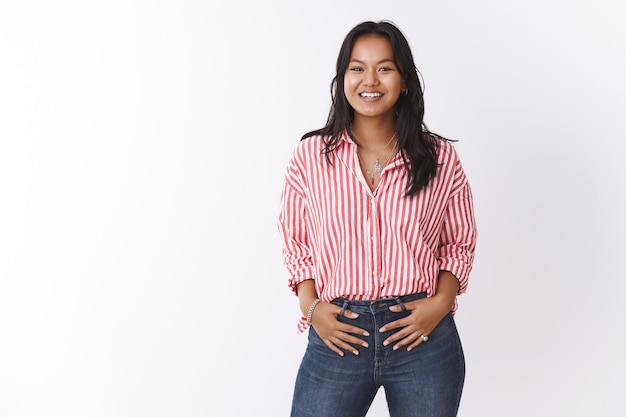 Colpo al coperto di bella giovane donna in camicetta a righe e jeans che tiene la mano fiduciosa in pantaloni e sorride alla macchina fotografica, sentendosi sicura di sé ed energica pronta per l'azione sul muro bianco