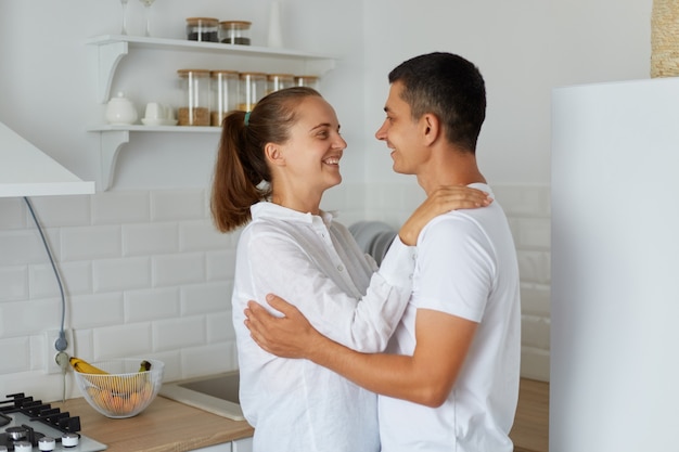 Colpo al coperto di donna sorridente che abbraccia con suo marito a casa in cucina leggera, coppia felice che sta insieme e si guarda l'un l'altro con amore e sentimenti romantici.