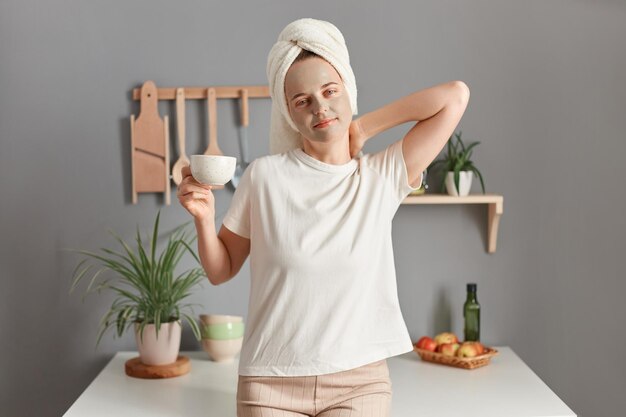 Colpo al coperto di giovane donna adulta attraente con maschera detergente sul viso in piedi in cucina a casa bevendo il suo caffè mattutino si sveglia presto