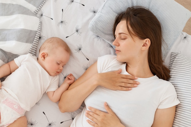 Colpo al coperto di giovane femmina adulta dai capelli scuri che indossa una maglietta casual bianca che dorme con la sua piccola figlia piccola, sdraiata sul letto a casa, felice momento di riposo.