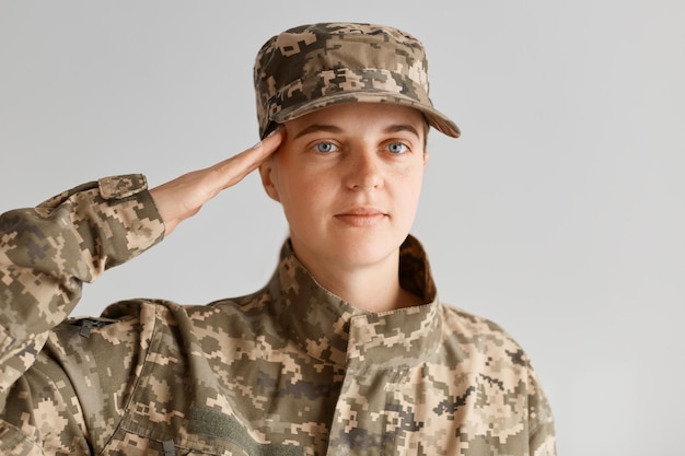 Colpo al coperto di giovane soldato dell'esercito donna attraente adulta che saluta, guardando la telecamera con un'espressione facciale seria, indossando l'uniforme militare, tenendo la mano vicino al tempio.