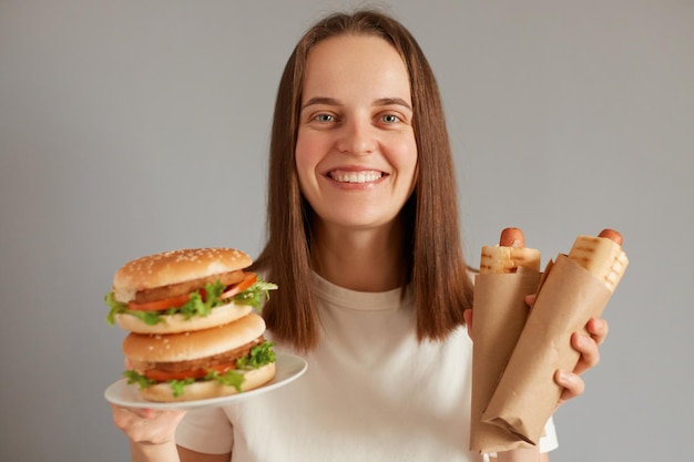 Colpo al coperto di sorridente allegra giovane donna adulta con i capelli scuri vestita con una maglietta bianca che tiene fuori un grande panino con hamburger e salsicce in pasta che ti dà uno spuntino fast food