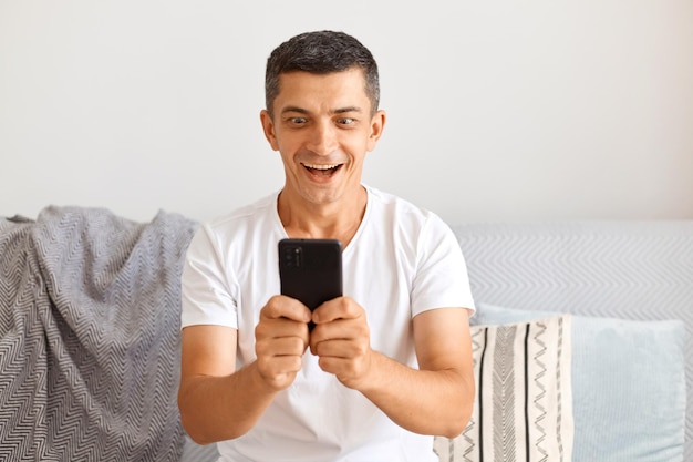 Colpo al coperto di un uomo dai capelli scuri estremamente felice che indossa una maglietta bianca seduto sul divano, con in mano il cellulare, guardando il display con grandi occhi, esprimendo eccitazione.