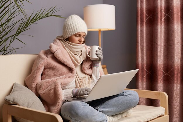 Colpo al coperto di una donna caucasica che indossa berretto e cappotto invernale seduta sul divano di casa lavorando online su notebook e bevendo tè caldo freelance lavora in appartamento freddo