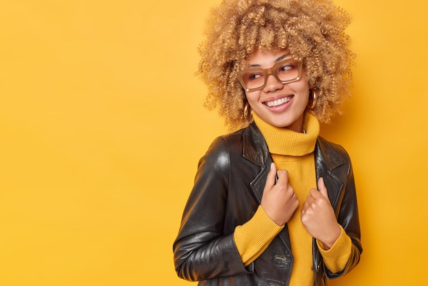 Colpo al coperto di una donna felice e gioiosa con i capelli ricci guarda con gioia qualcosa che è di buon umore vestito con un abito alla moda isolato su sfondo giallo, copia spazio per la tua promozione.