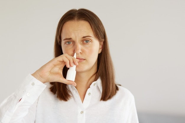 Colpo al coperto di una donna malata e accattivante con un aspetto piacevole che indossa una camicia bianca in stile casual, usando spray nasale, che soffre di naso che cola, guardando la telecamera con una faccia accigliata malata.