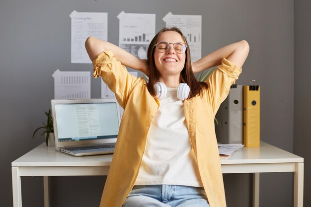 Colpo al coperto di una donna rilassata soddisfatta sorridente con i capelli castani che indossa una camicia gialla in posa in ufficio seduto vicino al tavolo con le braccia alzate e gli occhi chiusi che hanno una pausa gioendo alla fine del lavoro