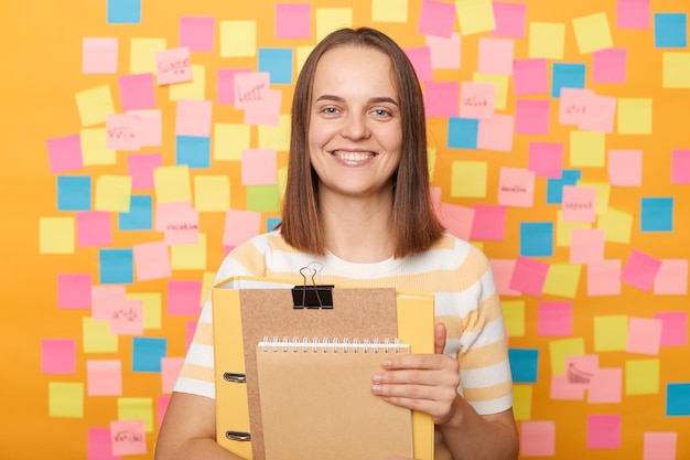 Colpo al coperto di una studentessa ottimista positiva con i capelli castani in maglietta a righe in piedi contro il muro giallo con schede memo che tengono documenti nella cartella di carta