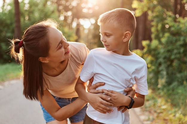 Colpo all'aperto della madre donna attraente che abbraccia il suo piccolo figlio che indossa una maglietta bianca, mamma che guarda il ragazzo con un sorriso, trascorrendo del tempo insieme nel parco estivo.