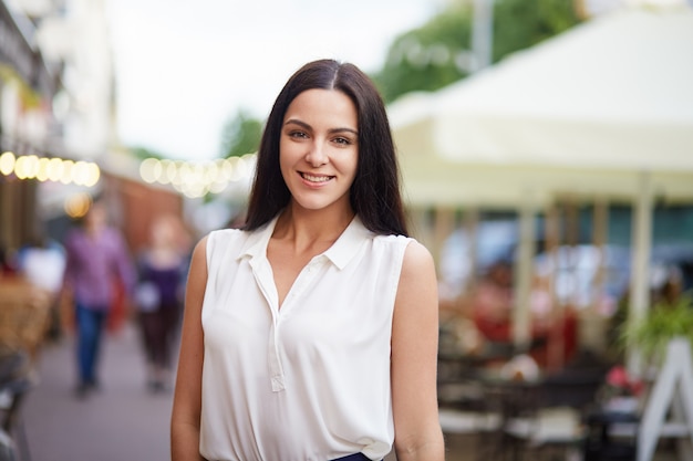 Colpo all'aperto di attraente giovane femmina bruna in camicia bianca, ha una passeggiata in città durante il giorno d'estate, pone alla macchina fotografica con un sorriso piacevole, ha un'espressione felice.